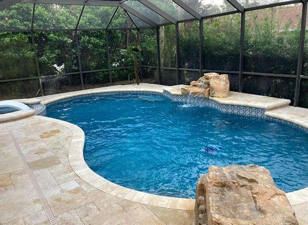 Swimming pool with a waterfall feature surrounded by a screened enclosure and light-colored stone.