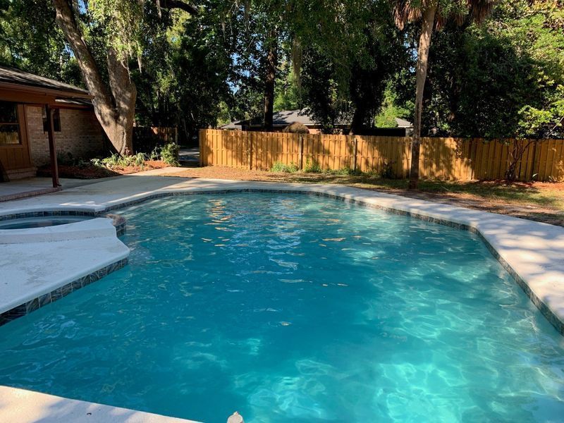 Swimming pool with blue water and surrounding concrete. Wooden fence in background. Sunny outdoor setting.