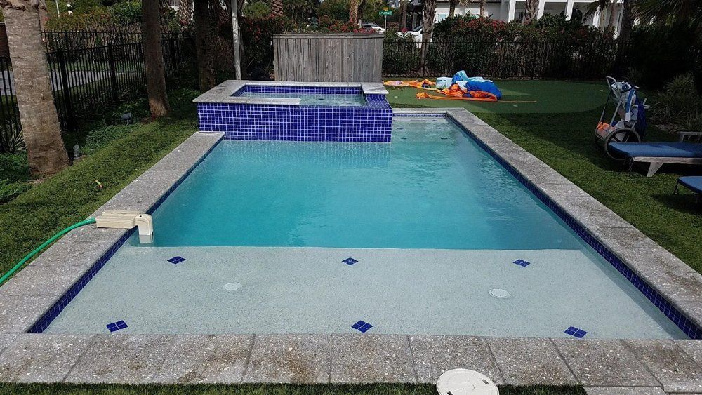 Rectangular swimming pool and small hot tub with blue tile accents, surrounded by stone and green grass.