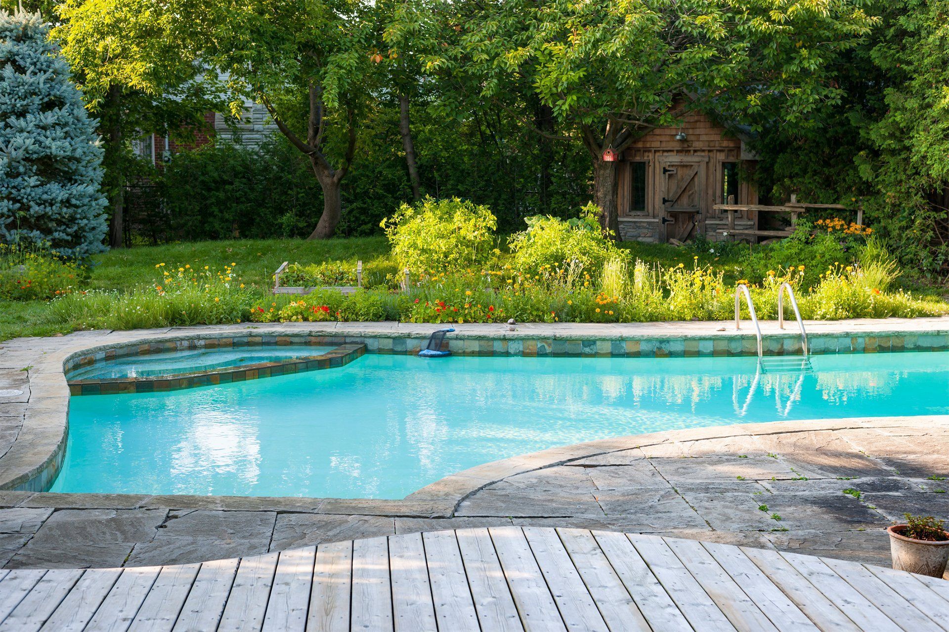 Swimming pool with surrounding stone patio, wooden deck, and a small wooden shed in a lush, green backyard. Swimming pool with surrounding stone patio, wooden deck, and a small wooden shed in a lush, green backyard.