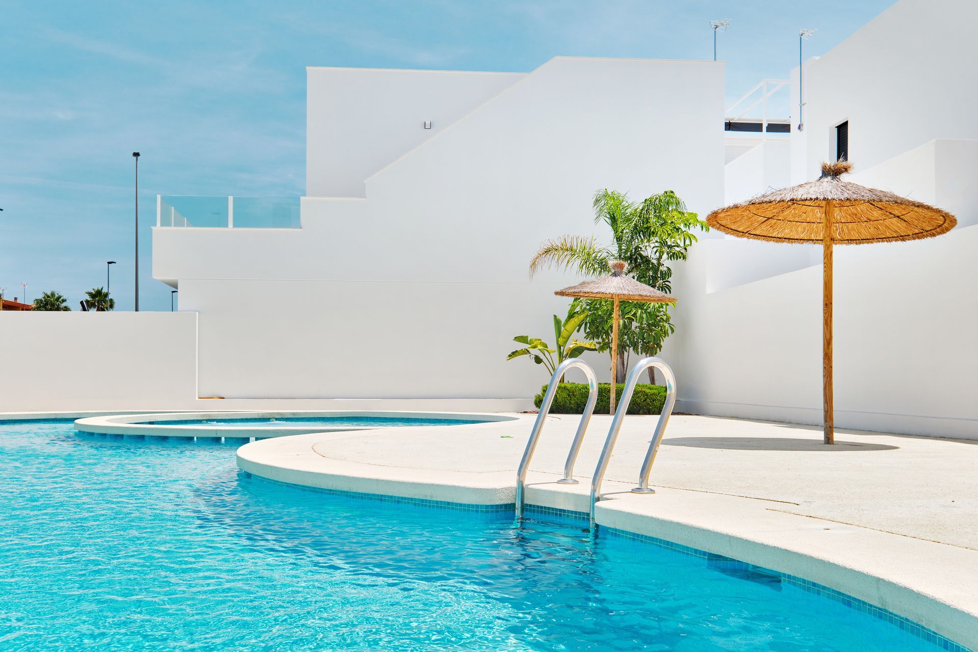 Pool with clear water and white building backdrop. Sunny day with straw umbrellas.