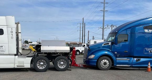 A blue semi truck is towing a white semi truck in a parking lot.