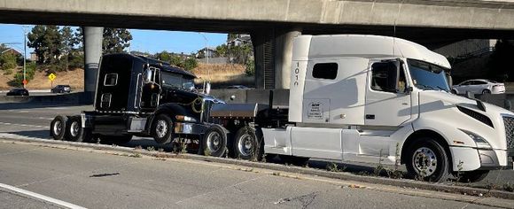 Two semi trucks are parked under a bridge on the side of the road.