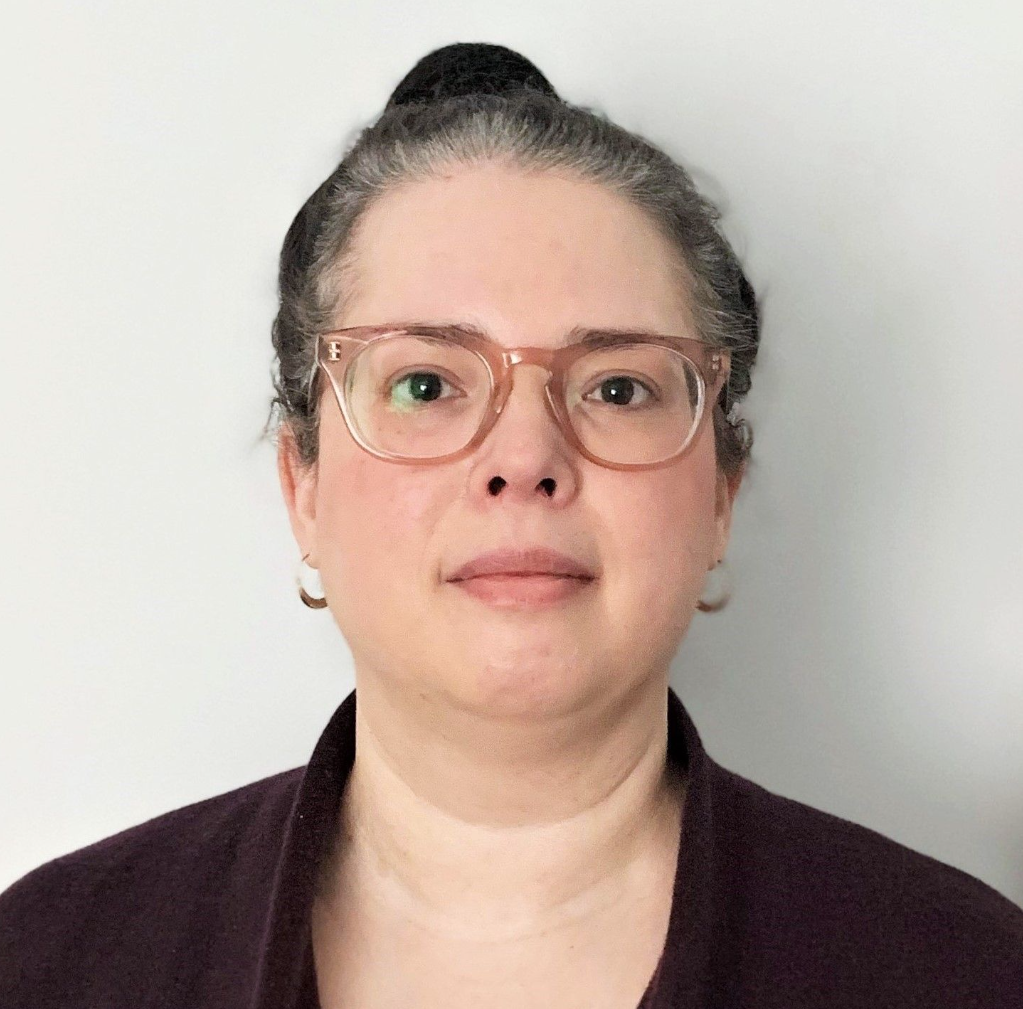 A woman wearing glasses and earrings is standing in front of a white wall.