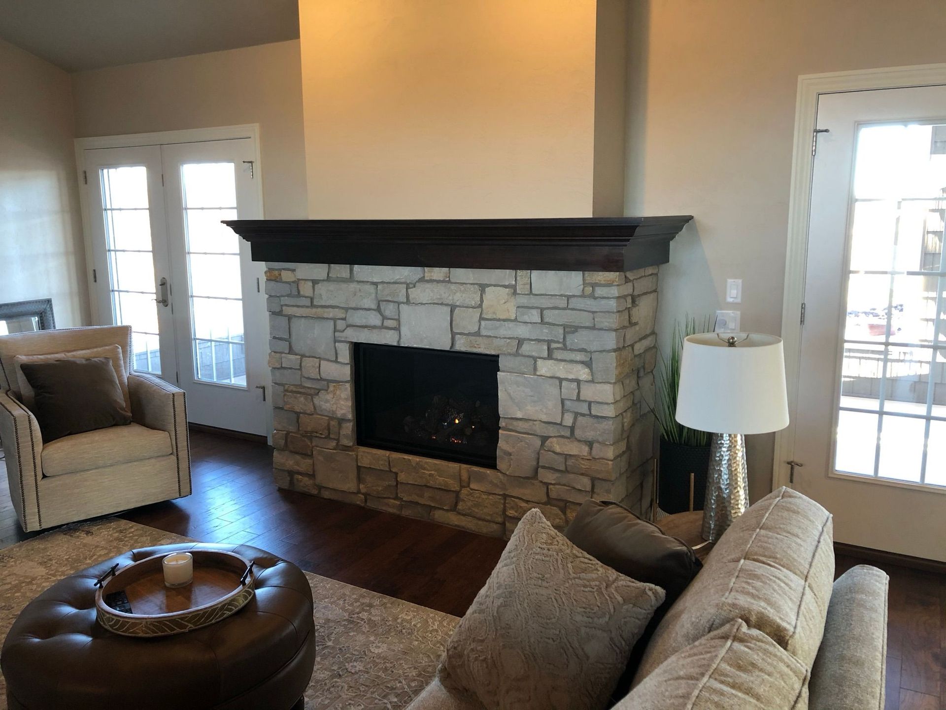 Stone fireplace in a living room with a dark wood mantel, flanked by windows. Beige walls.