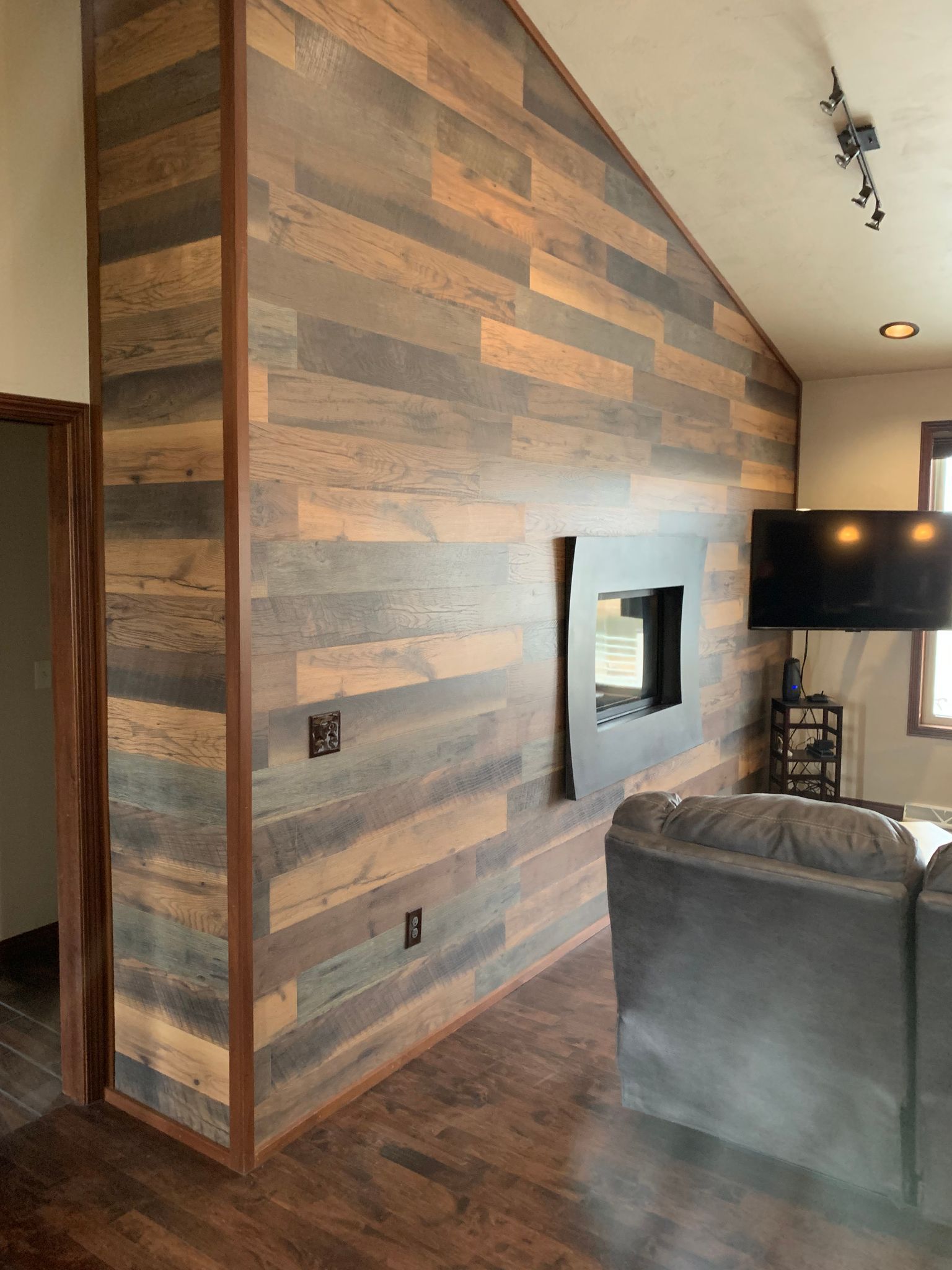 Wood-paneled accent wall with TV, neutral-toned wood flooring, and a section of a gray couch in a modern living space.