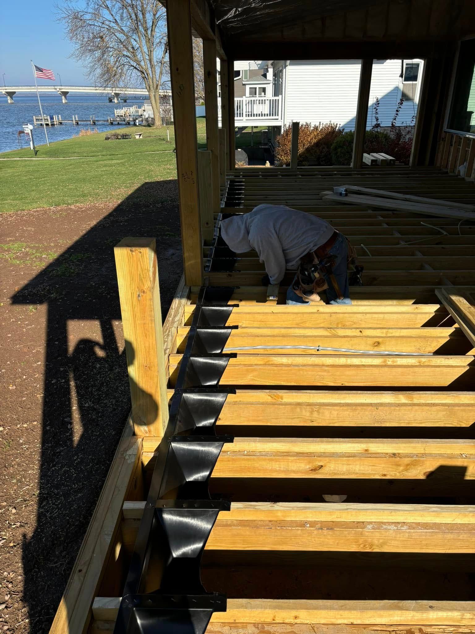 Construction worker installing deck boards on a waterfront structure. Wooden beams, blue sky, and water visible.