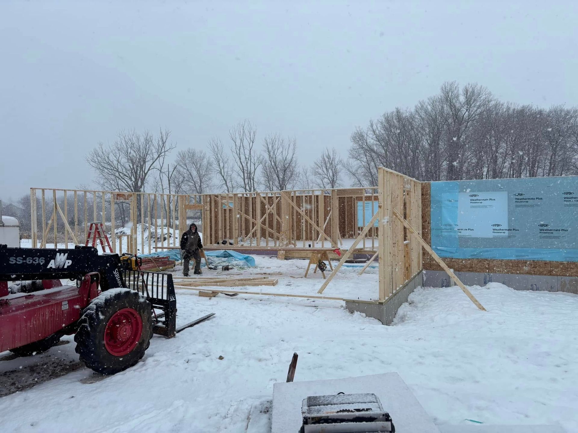 Construction site in winter, with a partially framed building and snow on the ground. A worker stands nearby.