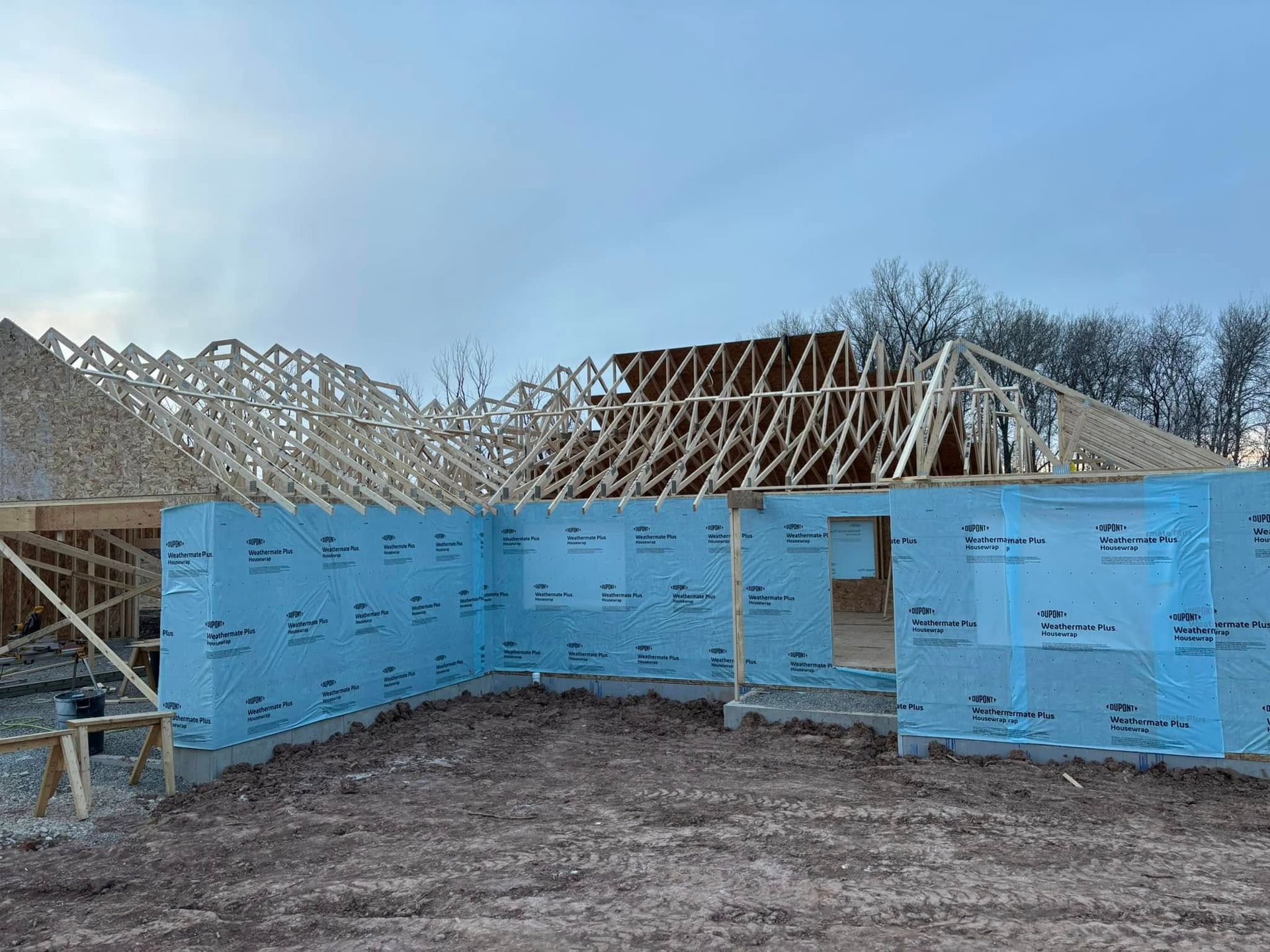 Construction site with wood framing, blue sheathing on walls, and trusses under a cloudy sky.