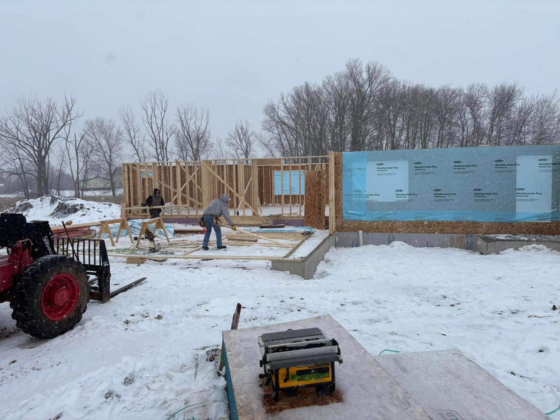 Construction workers building a house frame in the snow; tractor and saw visible on a snowy lot.