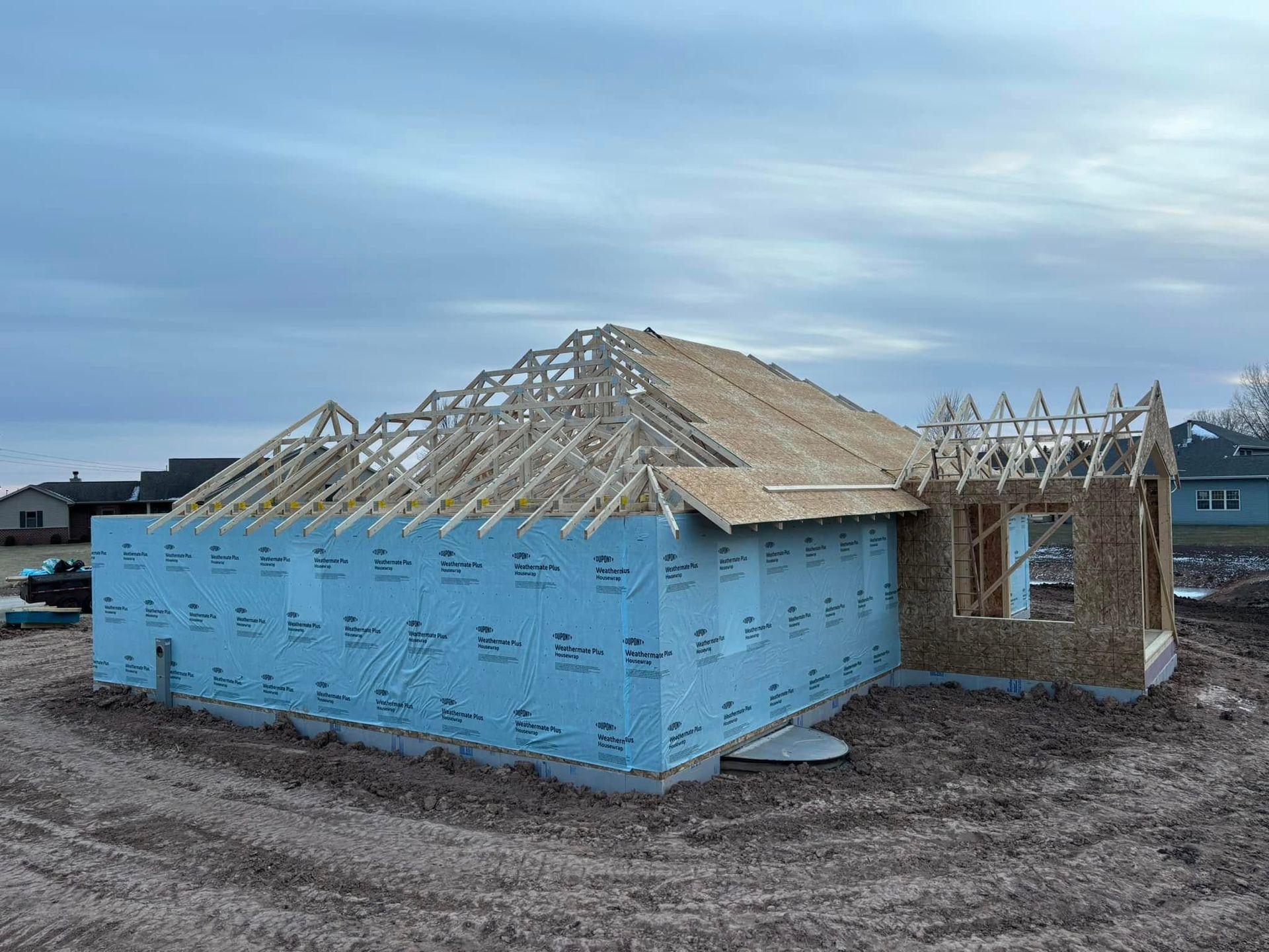 House under construction, framed roof, blue wrap on exterior walls, overcast sky.