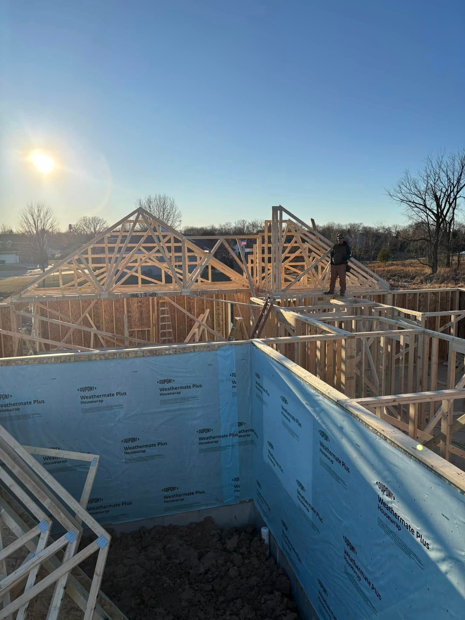 Construction site with a partially framed house. A worker stands atop the roof frame in the sun.