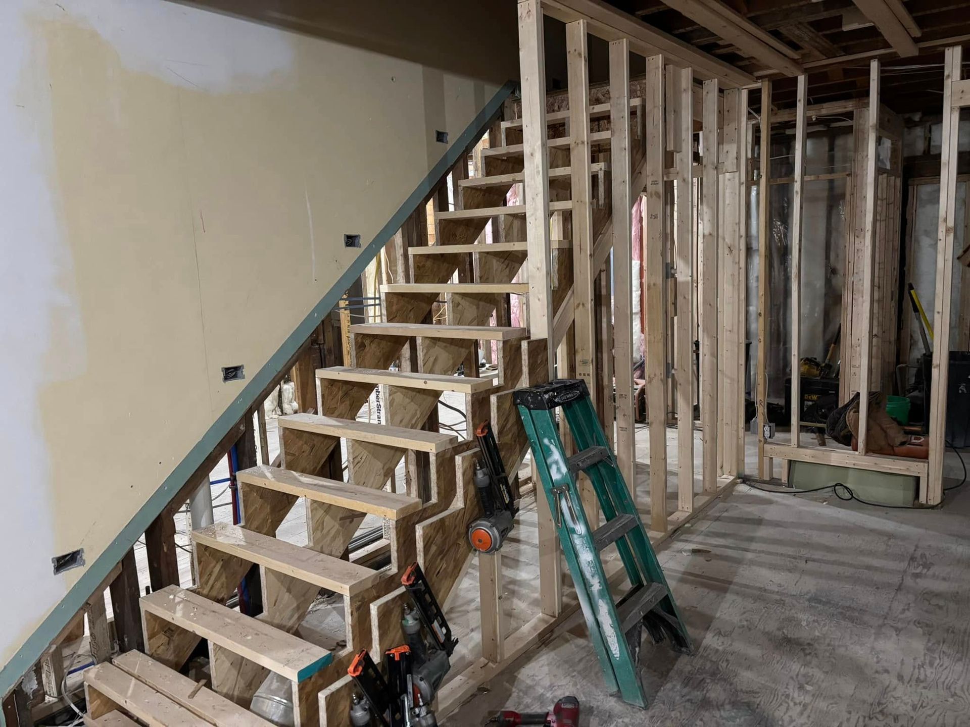 Wooden staircase under construction in a room with framed walls; a green ladder leans against the wall.