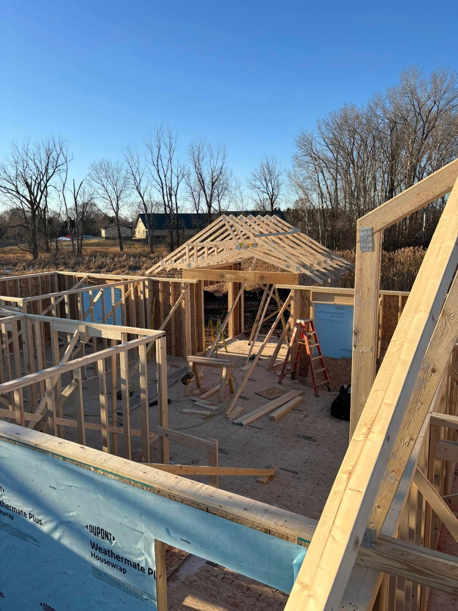 Construction site: wooden house frame, partially built, with a completed gazebo roof under a clear blue sky.