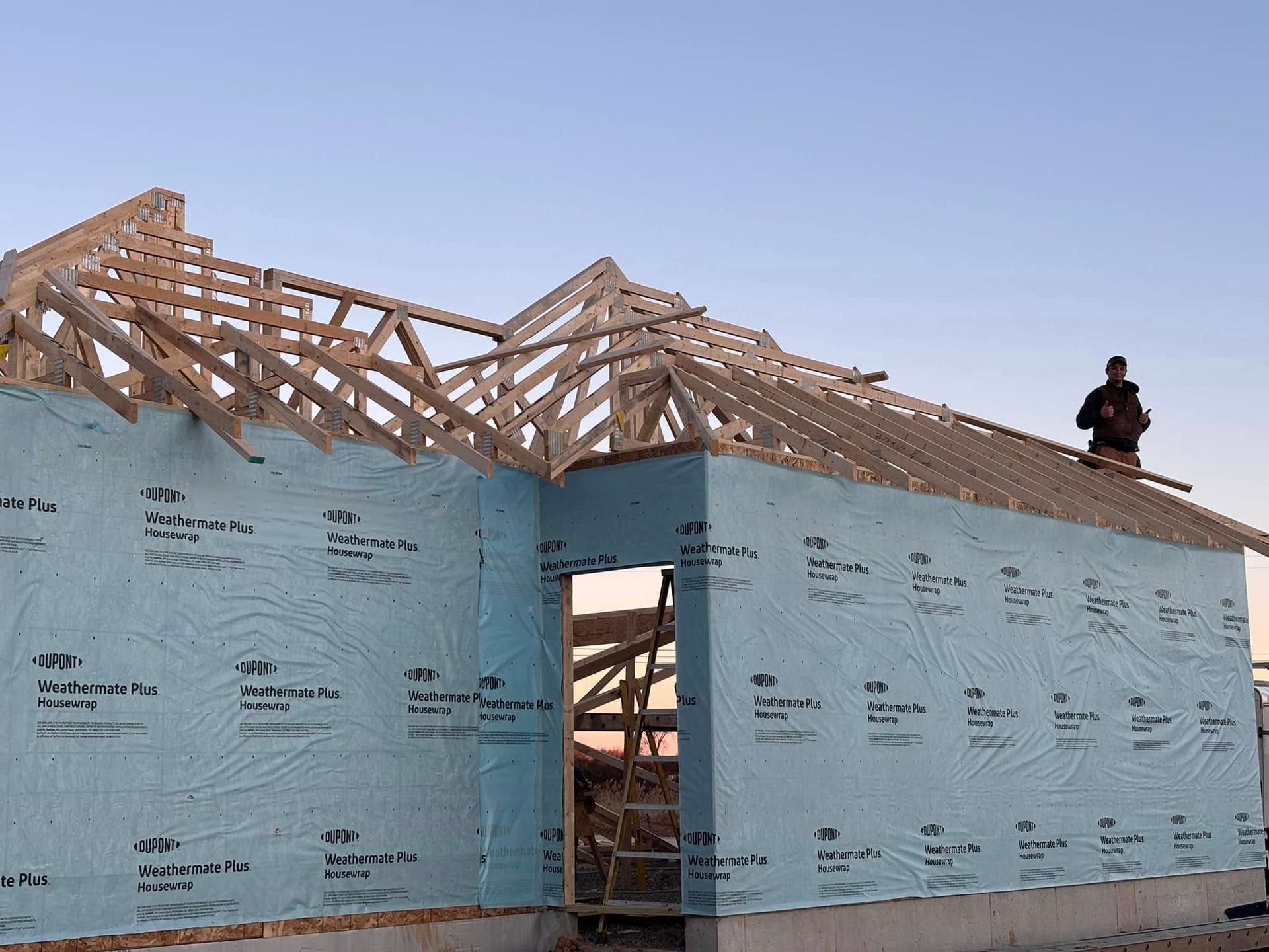 Construction site: House frame with wooden trusses. Person on the roof. Blue sheathing on walls, setting sun.