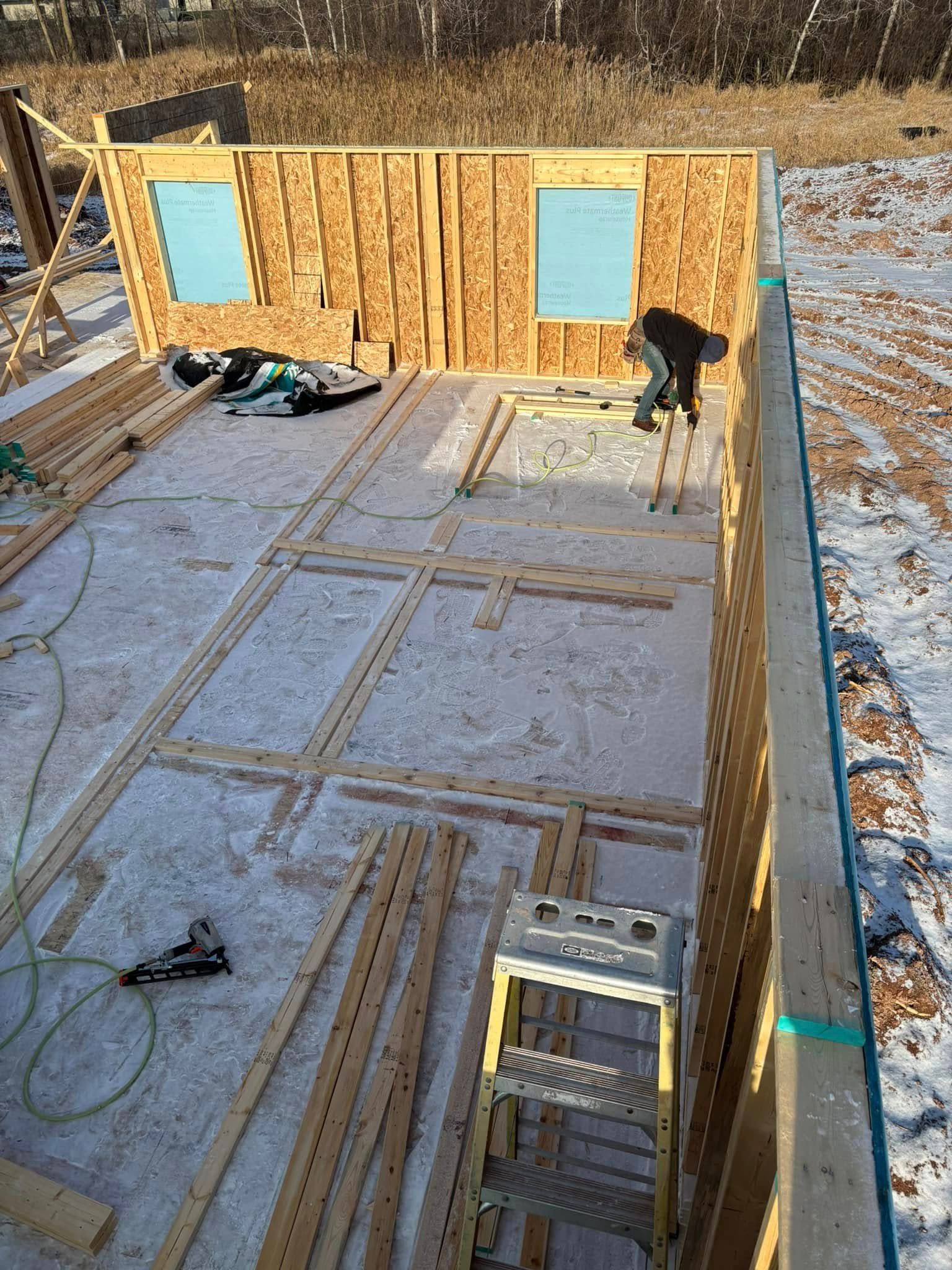 Construction site with wooden frame walls, a worker, and a ladder. Snow on the floor.