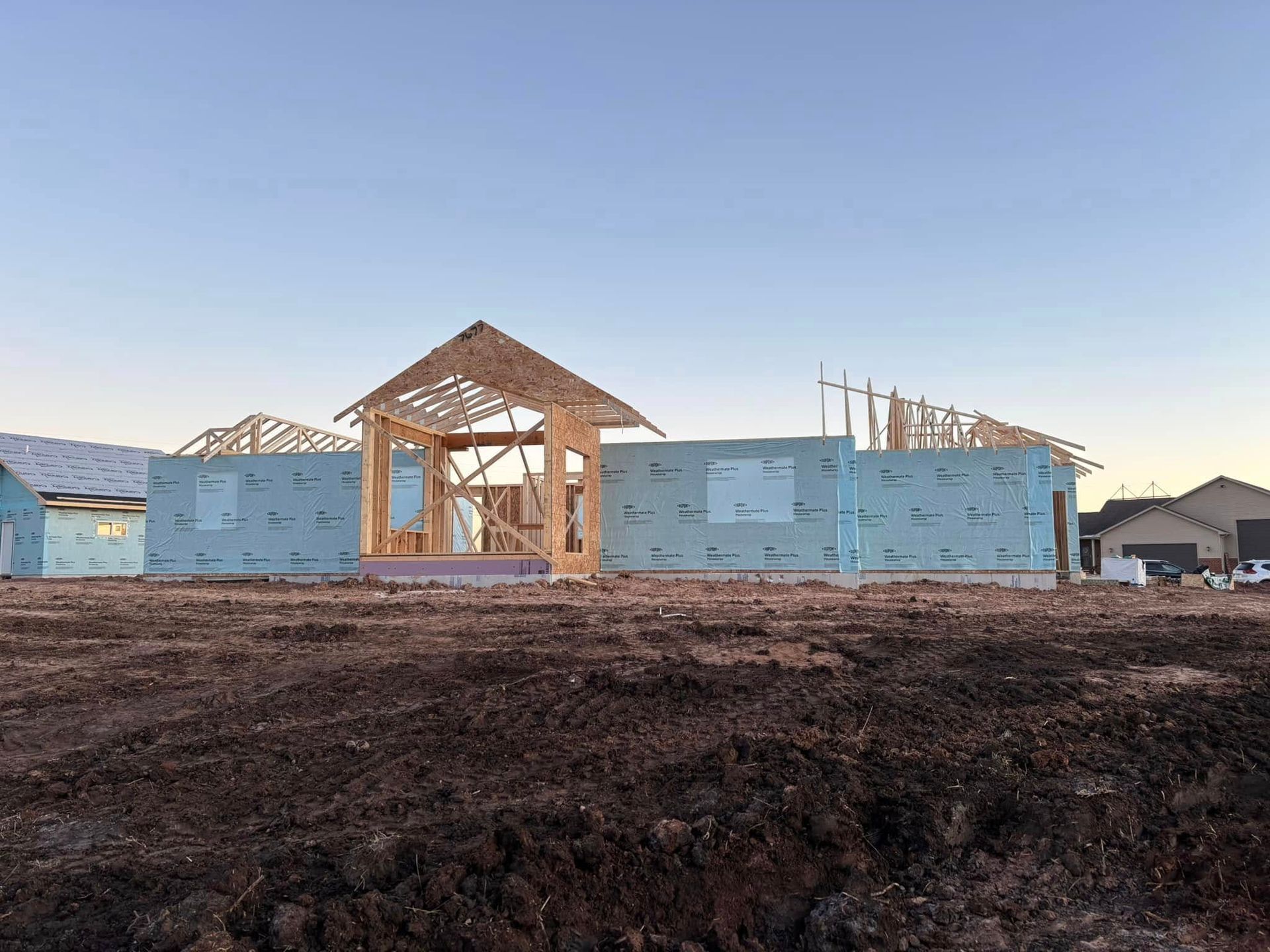 Construction site: Wood frame house under construction, blue wrap, dirt ground, clear sky.