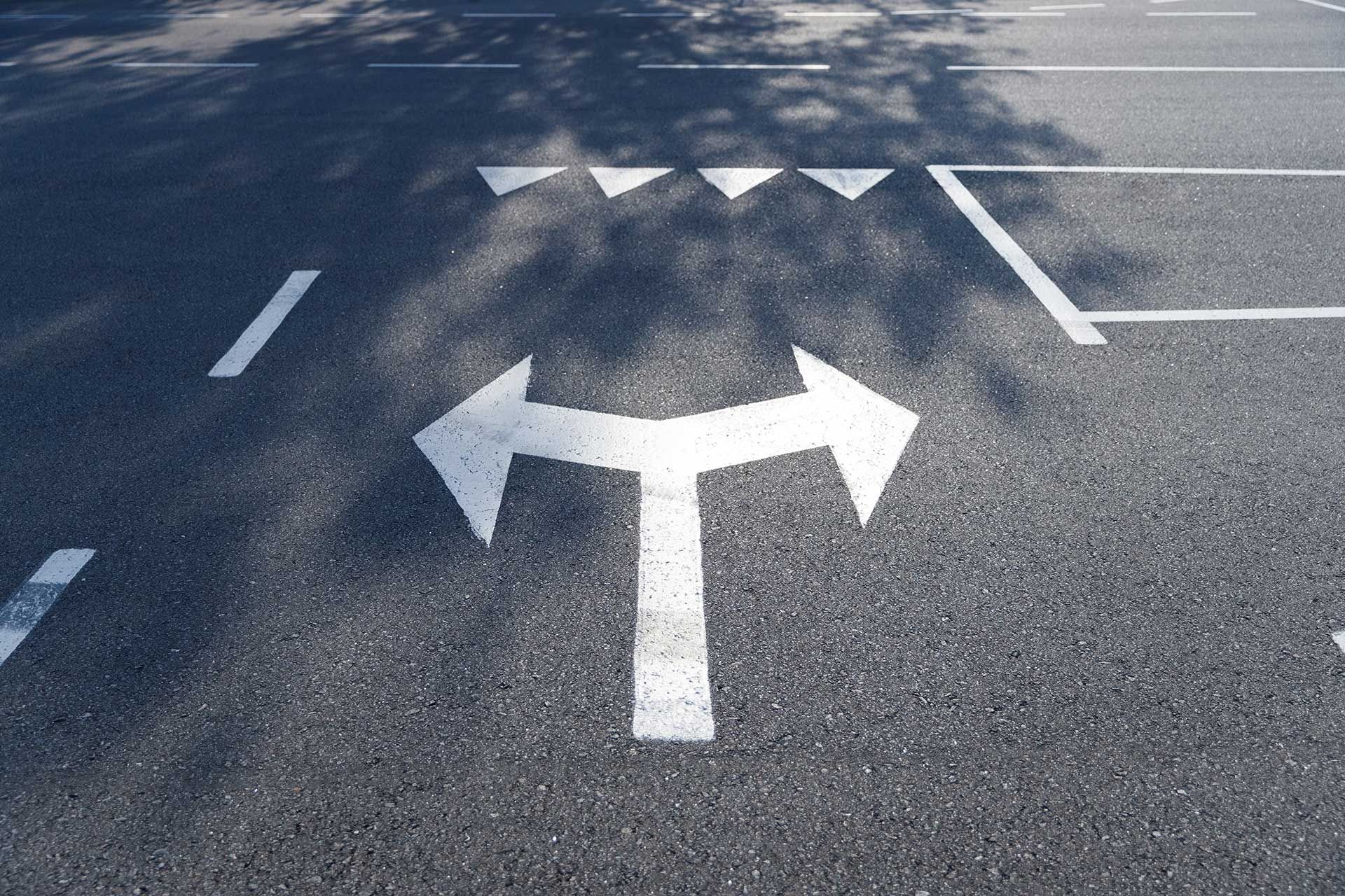 A white road marking showing a T-junction split with two arrows pointing left and right on grey asphalt.
