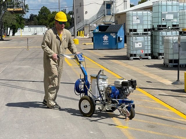 A worker wearing a yellow hard hat and beige jumpsuit operates a blue industrial paint striper on a paved outdoor lot.