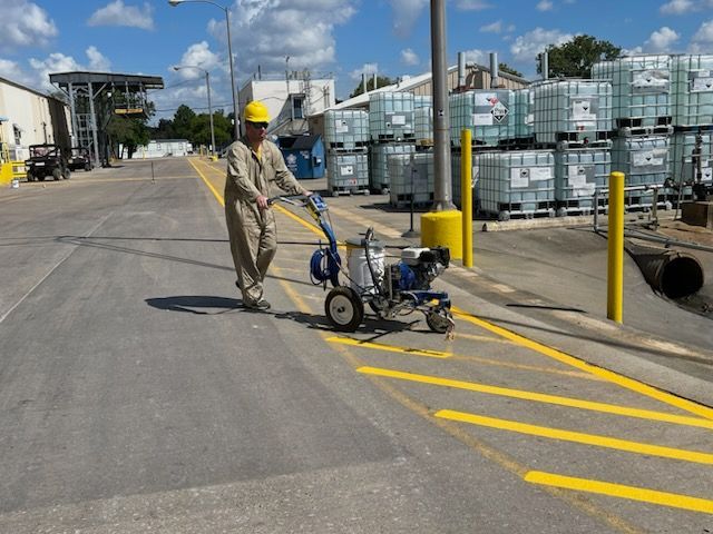 A person in protective work gear uses a mechanical sprayer to paint yellow stripes on an asphalt parking lot.