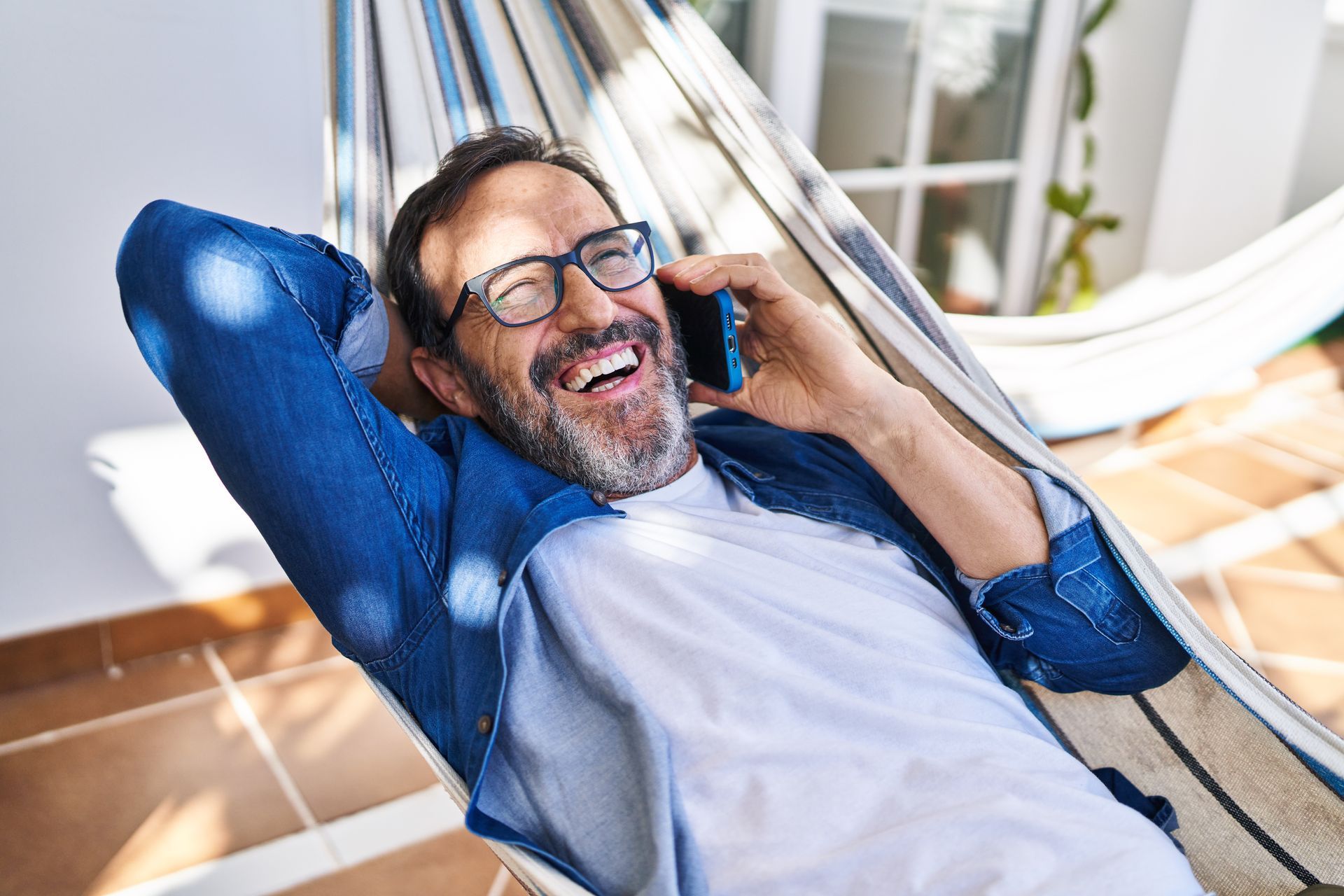 Man in glasses laughing on a hammock while talking on a phone; outdoors.