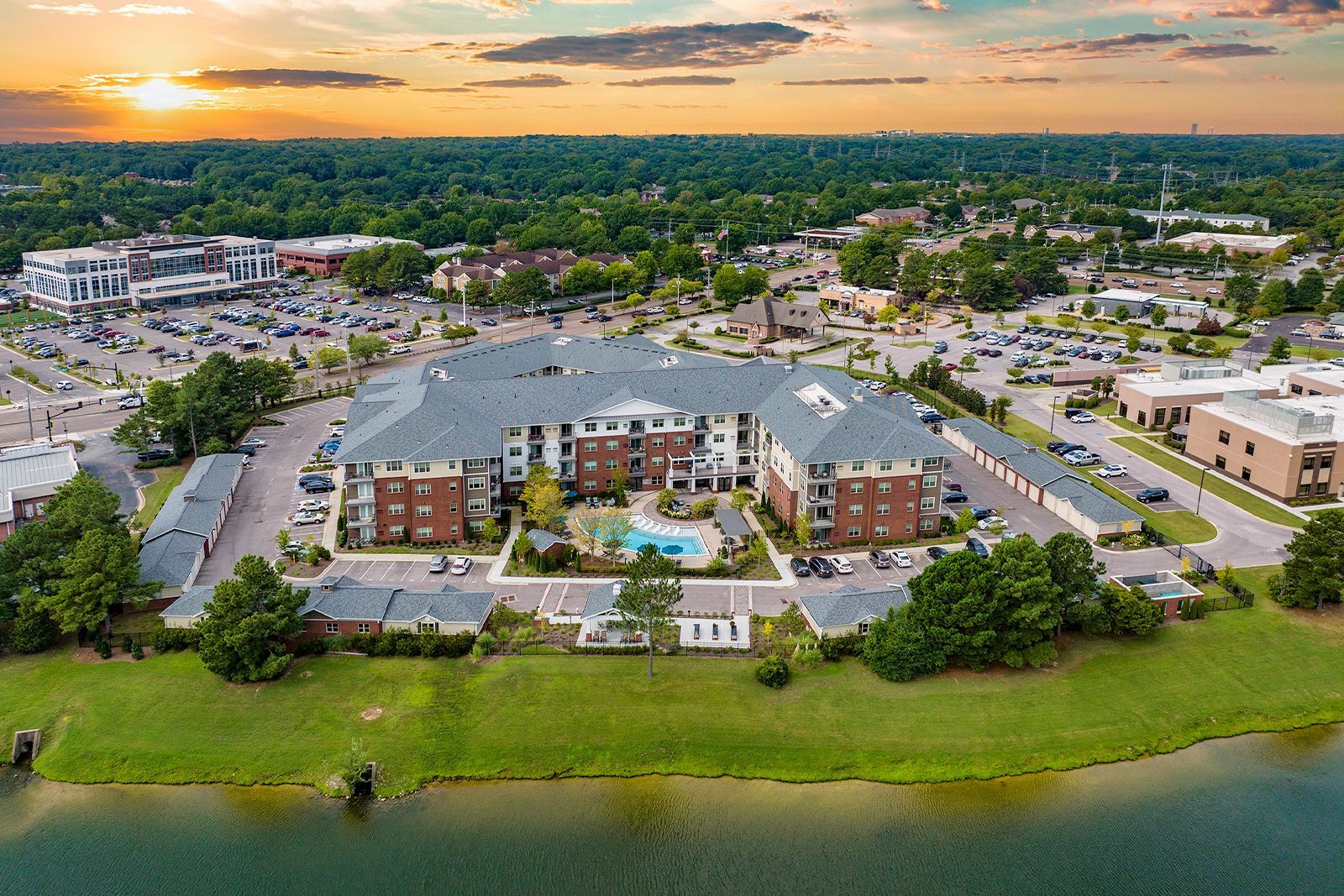 Aerial view of apartment buildings with a pool, surrounded by a parking lot and a green landscape near a lake.