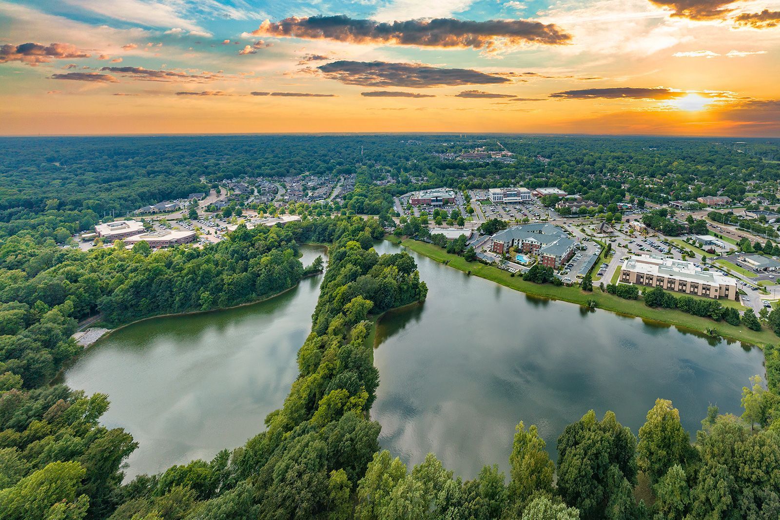 Aerial view of a lake surrounded by trees and a suburban town at sunset; orange and blue hues in the sky.