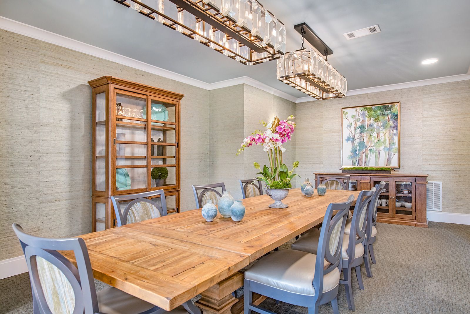 Dining room with a long wooden table, chairs, china cabinet, and chandeliers.