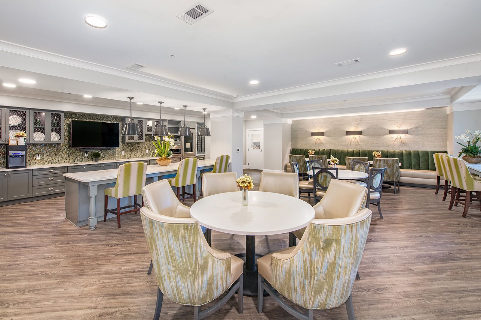 Dining area in senior living facility. Round tables and upholstered chairs. Gray cabinetry, hardwood floors.