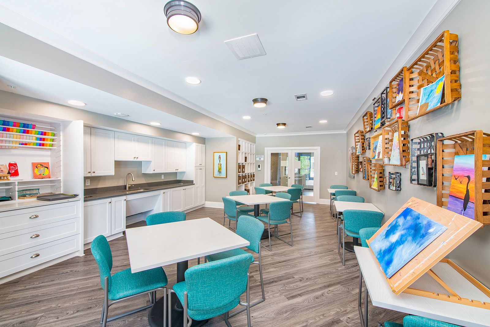 Art studio with white tables, turquoise chairs, paintings, and art supplies.