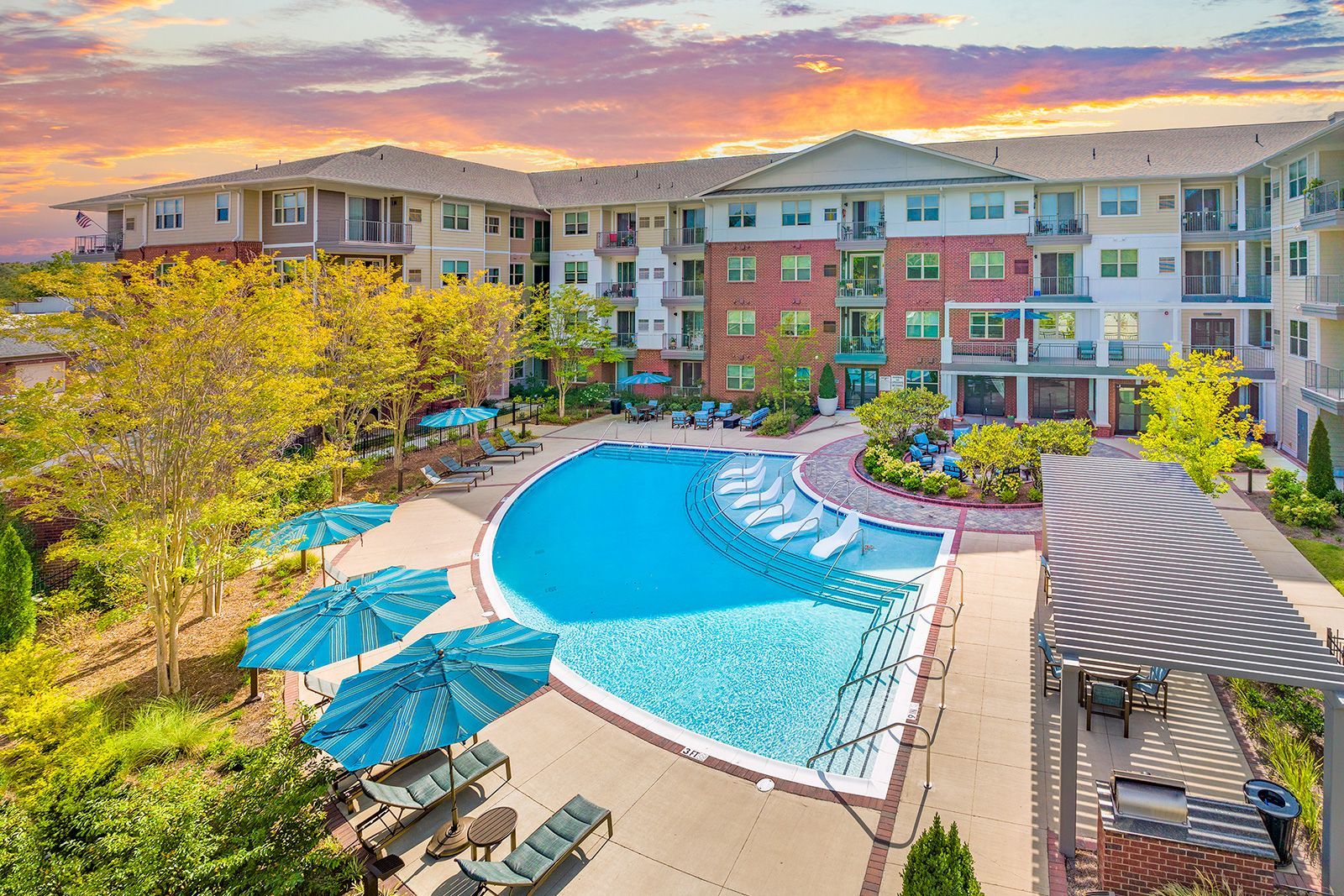 Apartment complex with pool, lounge chairs, umbrellas, and landscaping. Sunset sky.