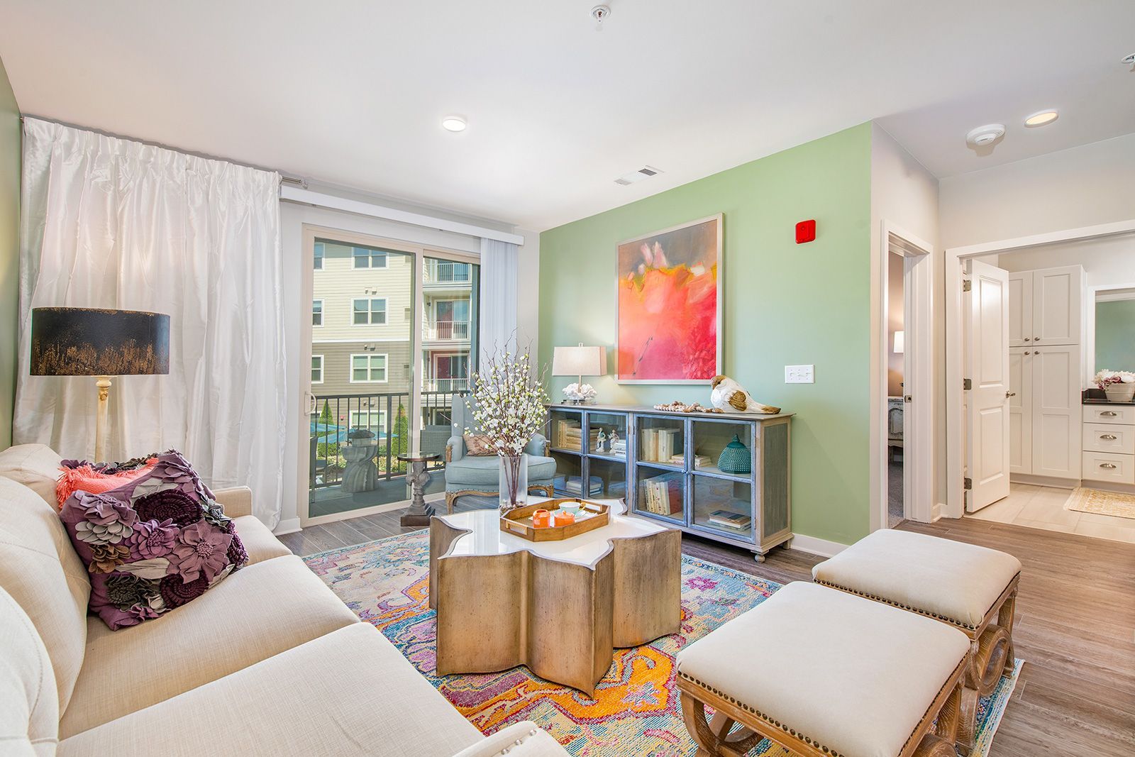 Cozy living room with sofa, colorful rug, and open door to a balcony. Mint green walls, artwork, and neutral furniture.