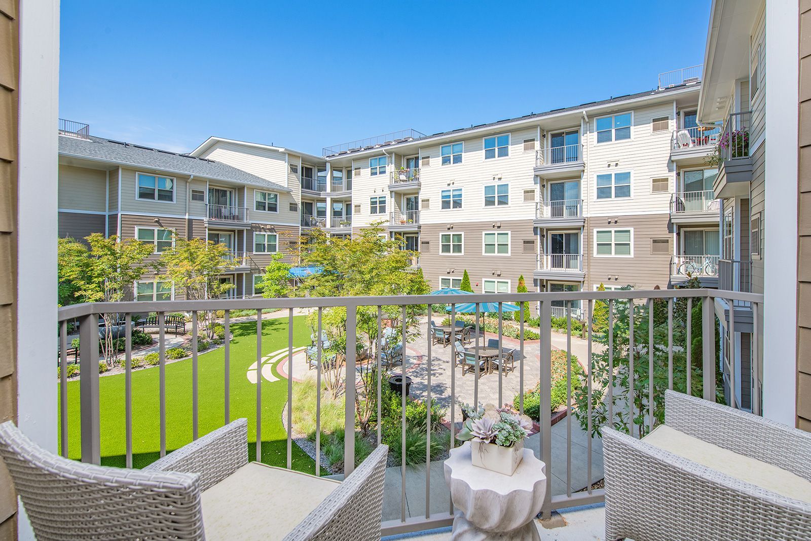 Balcony view of a courtyard with chairs, green grass, trees, and multi-story buildings in background.