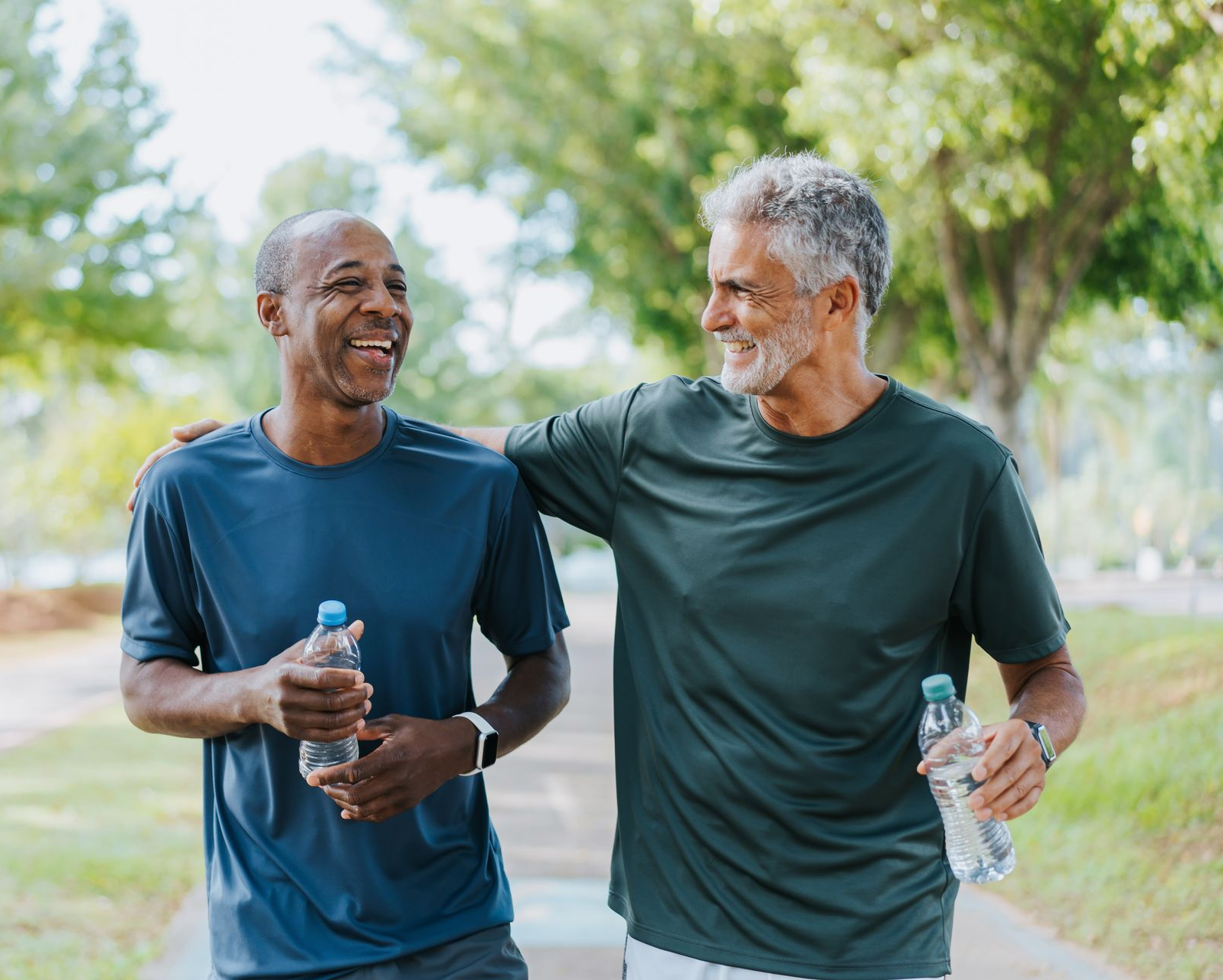 Two men walking on a path, smiling, with arms around each other. Holding water bottles, in a park.