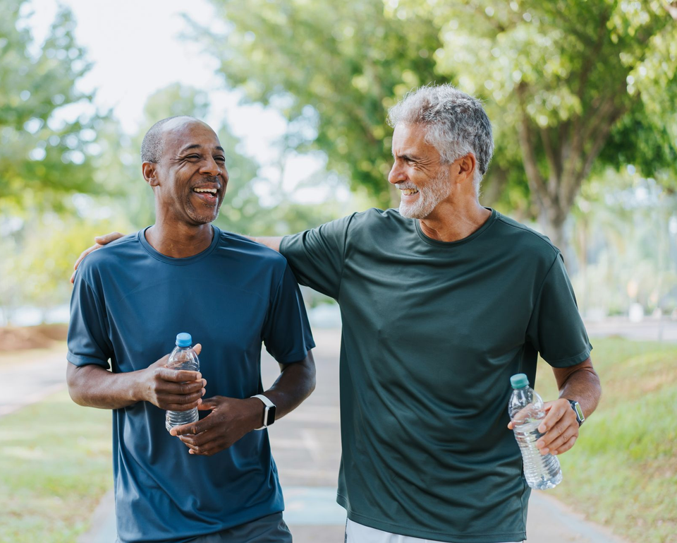 Two men walking on a path, smiling, with arms around each other. Holding water bottles, in a park.