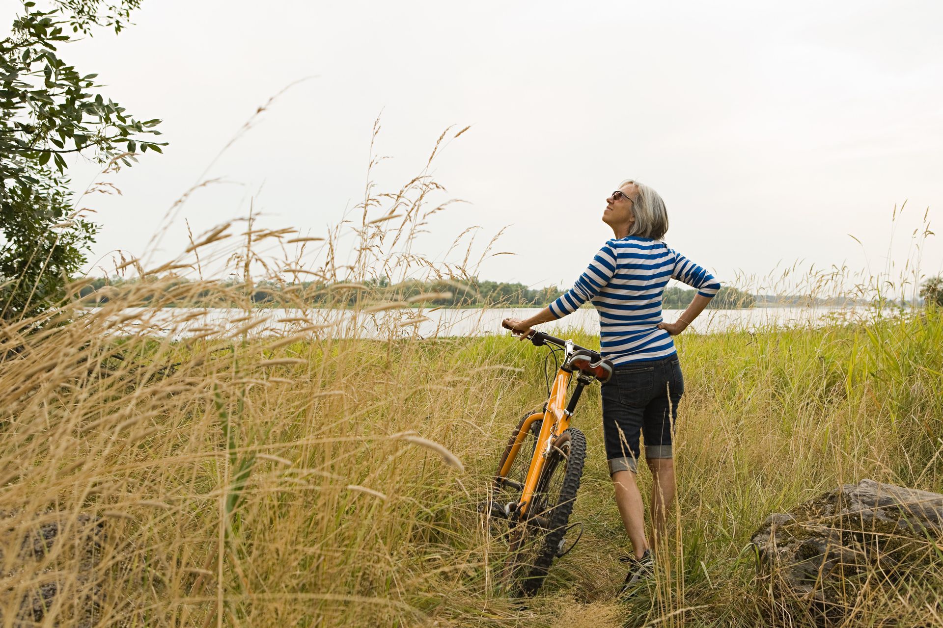 Woman with bike looks up at sky, standing in tall grass near a lake.