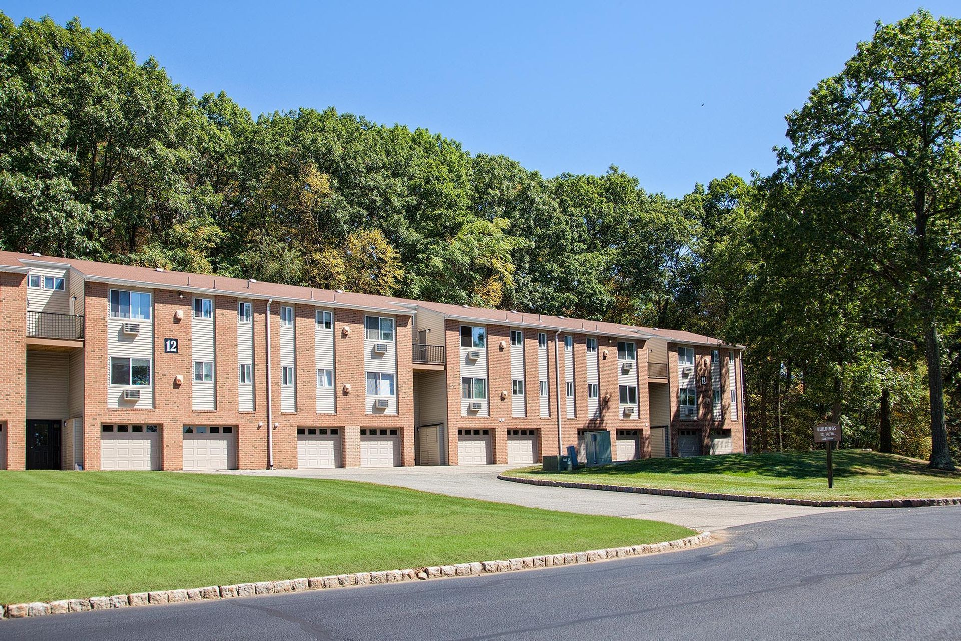 Exterior view of a brick apartment building with garages, green lawn, and trees.