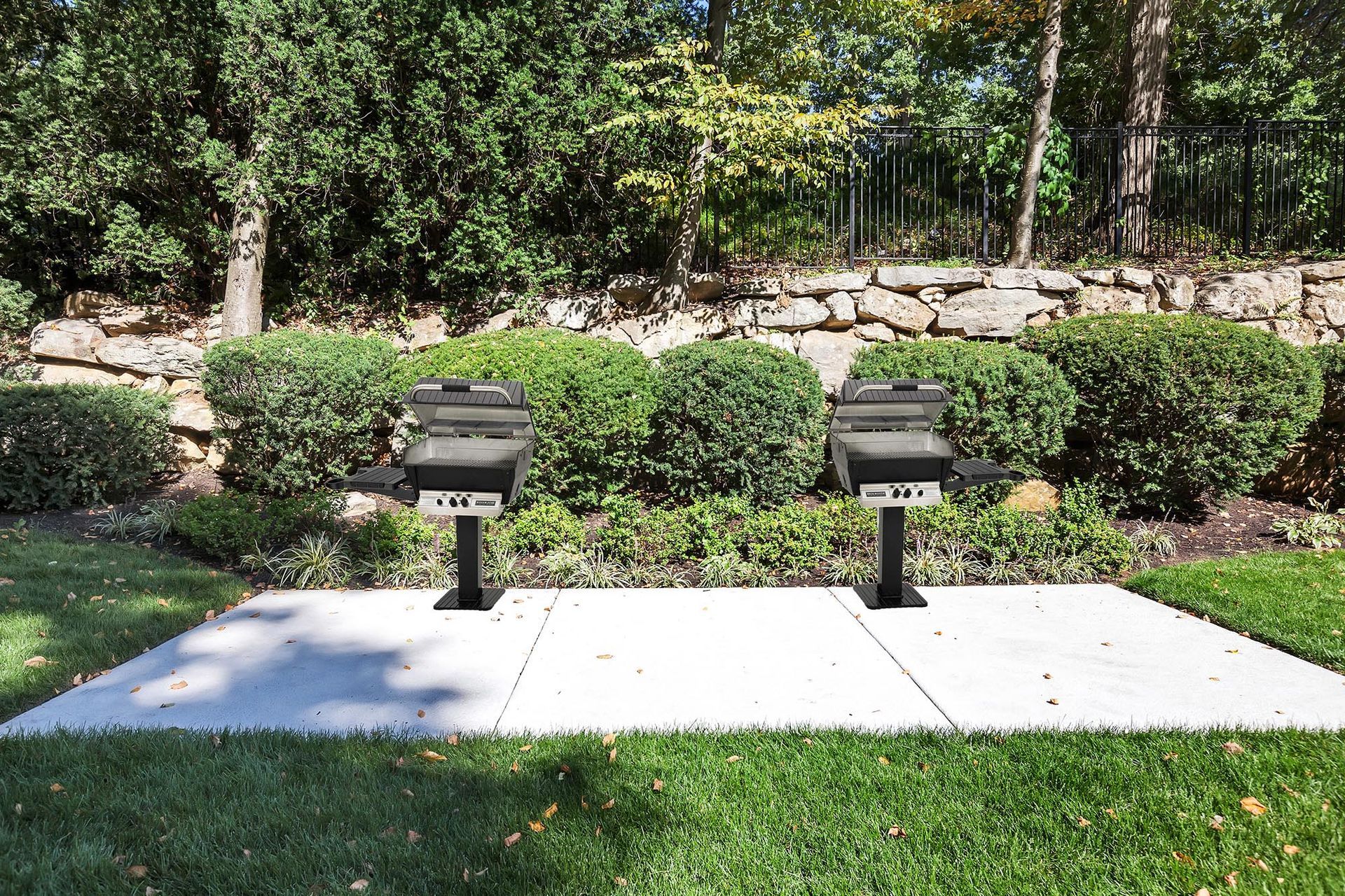 Two outdoor grills on a concrete pad in a landscaped communal area.