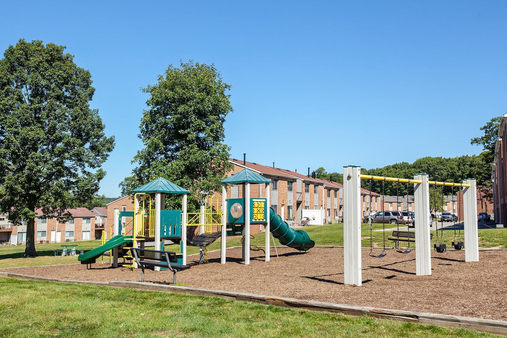Playground with slides and swings at an apartment complex, with trees and townhomes in the background.