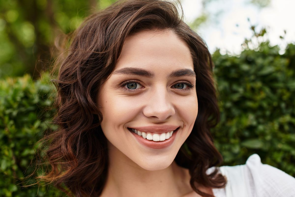 Smiling woman with wavy brown hair outdoors against a green leafy background