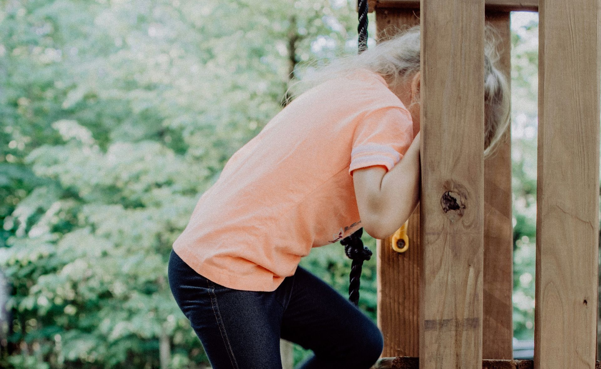 Child wearing a peach shirt and jeans hides behind a wooden structure outdoors.