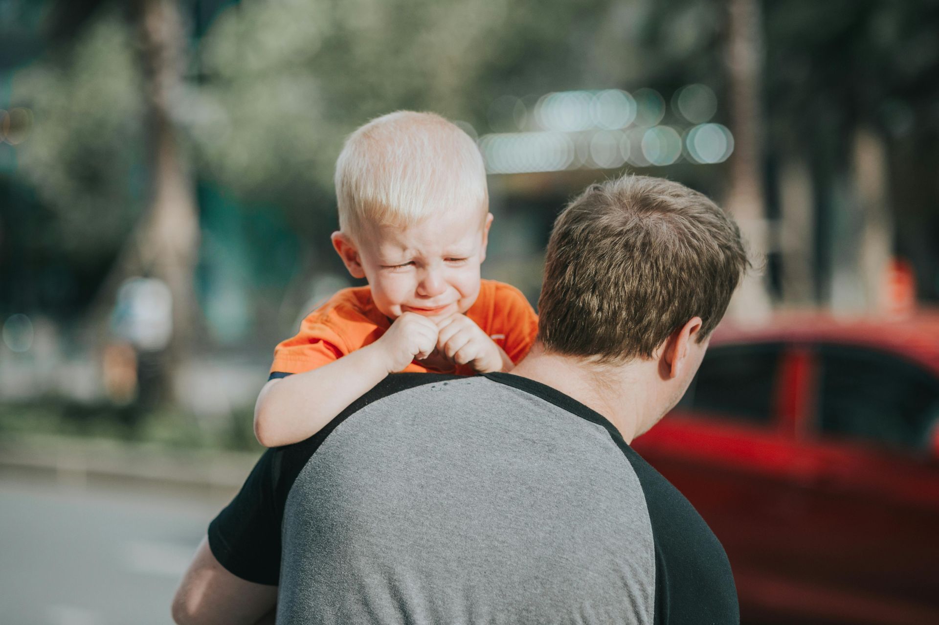 Child crying on a person's shoulders, outside. The person wears gray shirt. Red car and trees in background.