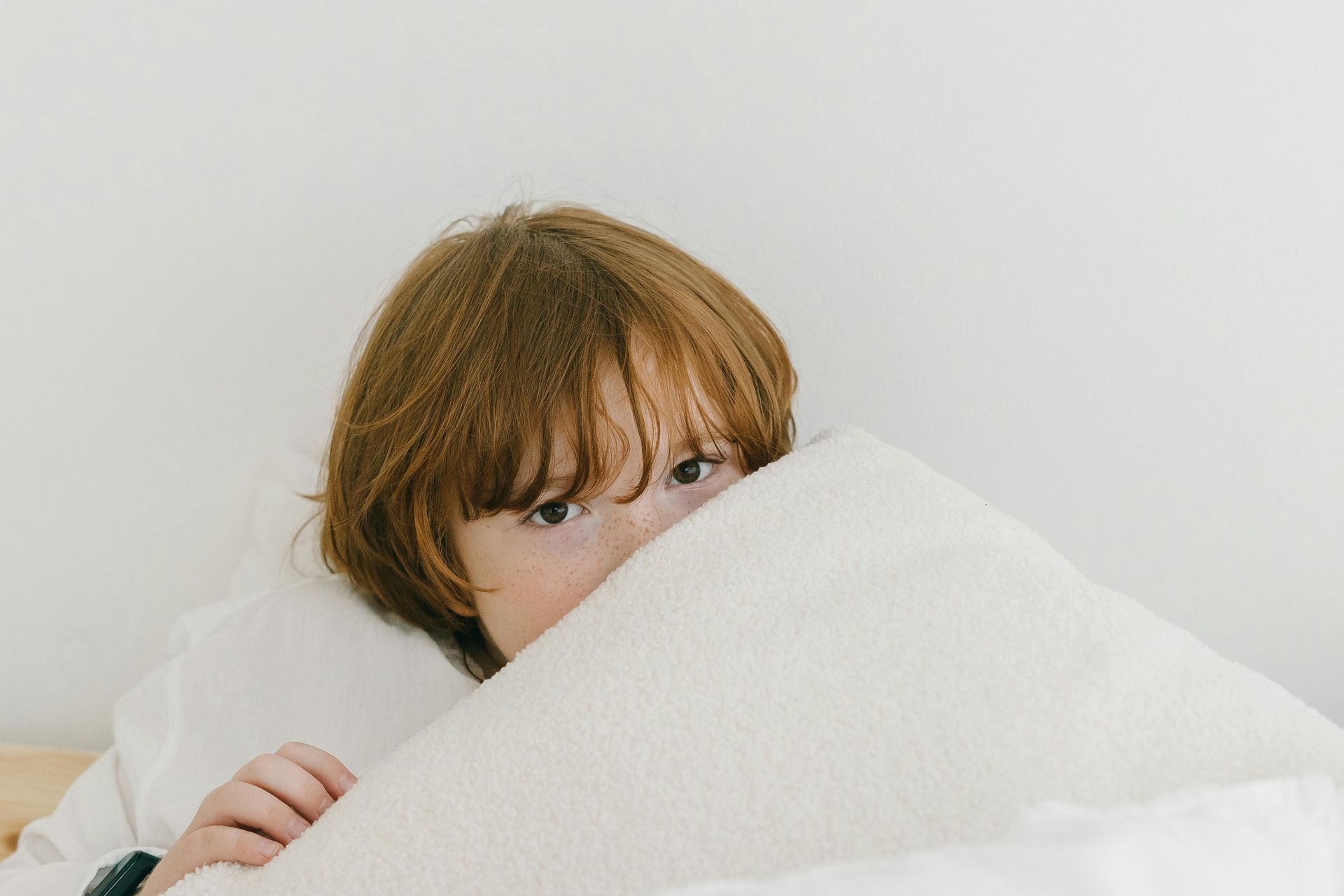 Child with reddish-brown hair peeks over a white pillow on a bed, eyes wide.
