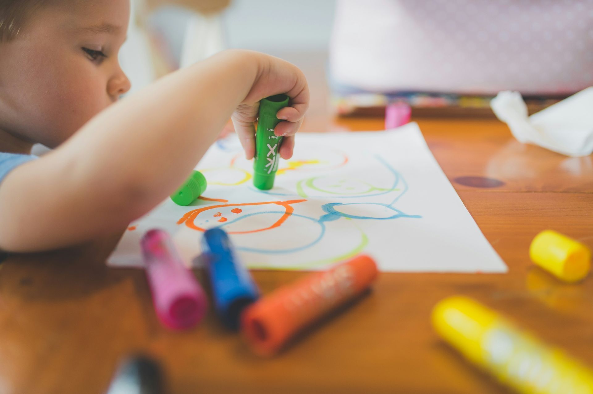 Child coloring with crayons on a white paper on a wooden table; scattered crayons.