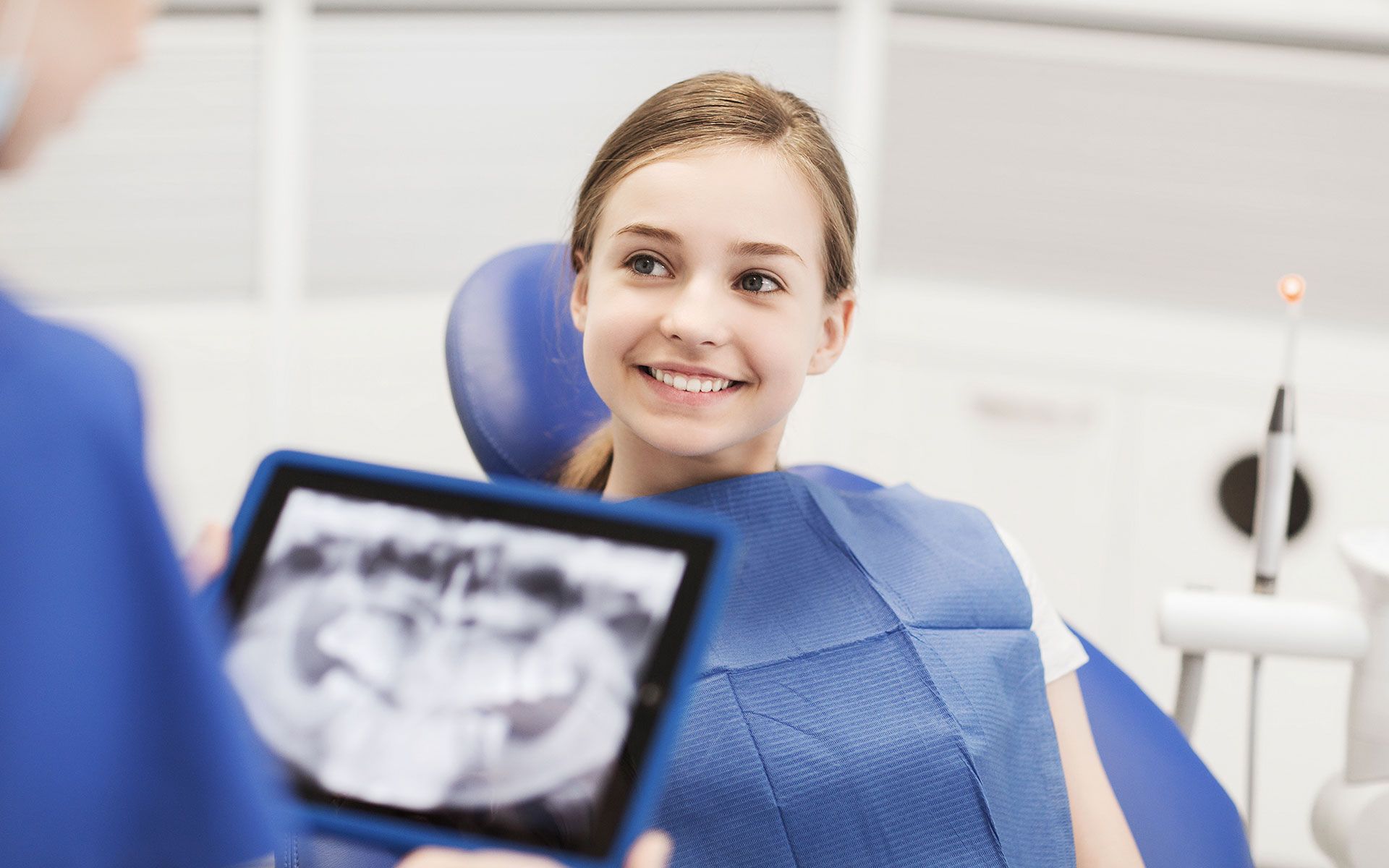 A young girl is sitting in a dental chair while a dentist shows her an x-ray of her teeth.