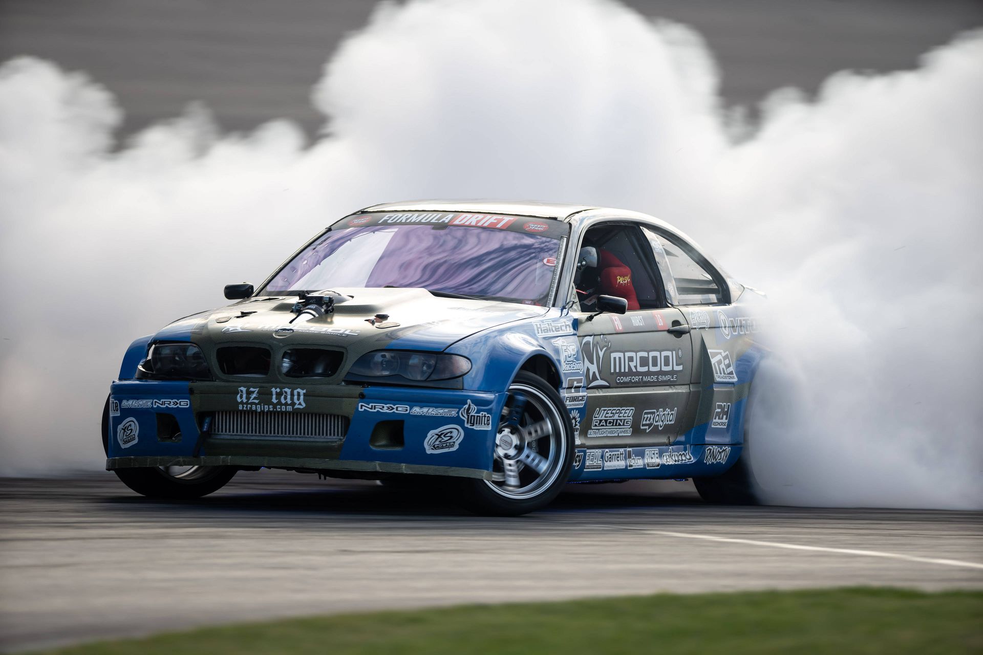 A blue and white bmw is drifting on a track with smoke coming out of the tires.