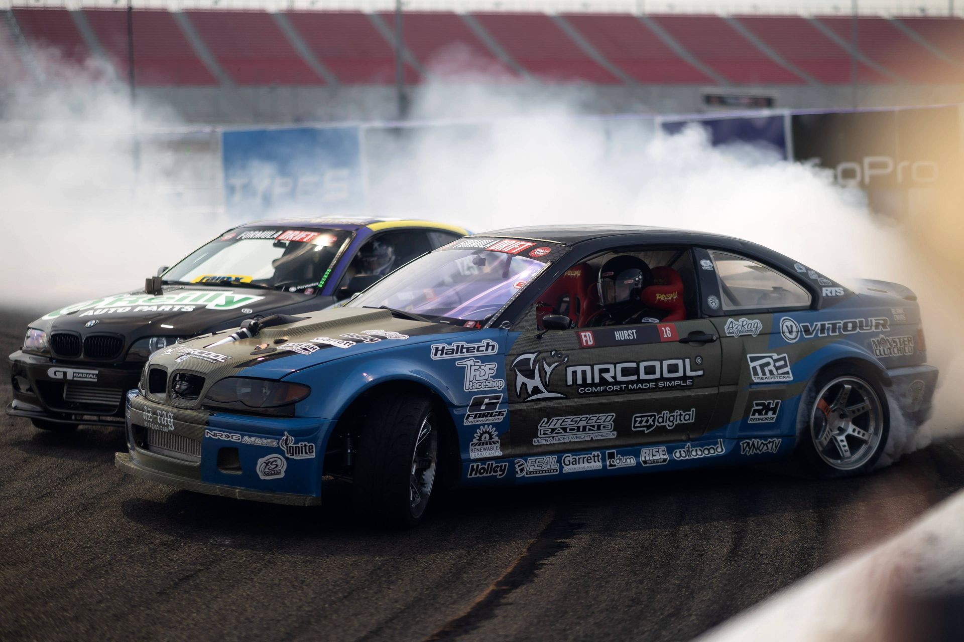 Two cars are drifting on a track and one is sponsored by mr cool