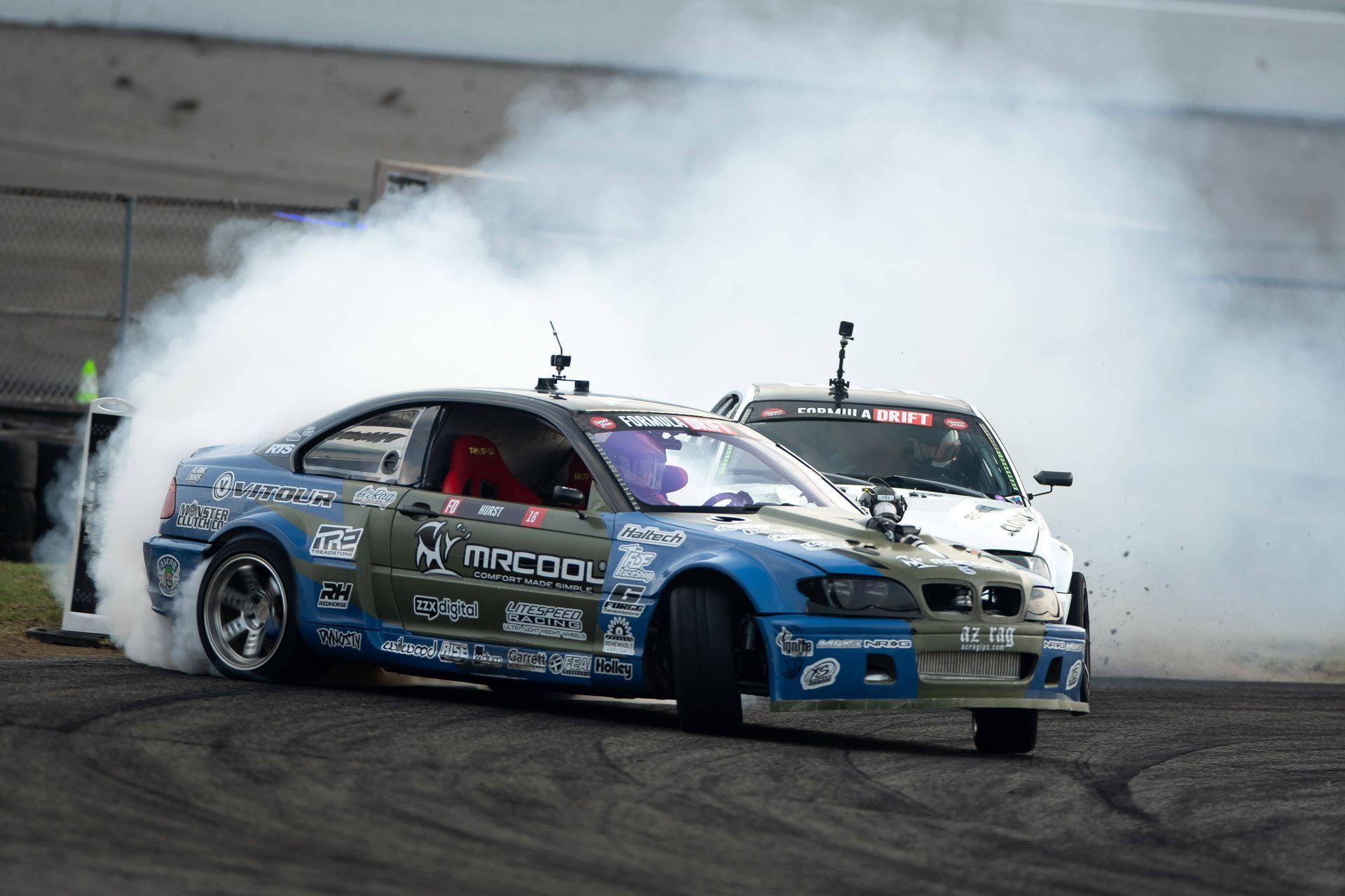 Two cars are drifting on a track with smoke coming out of the tires.