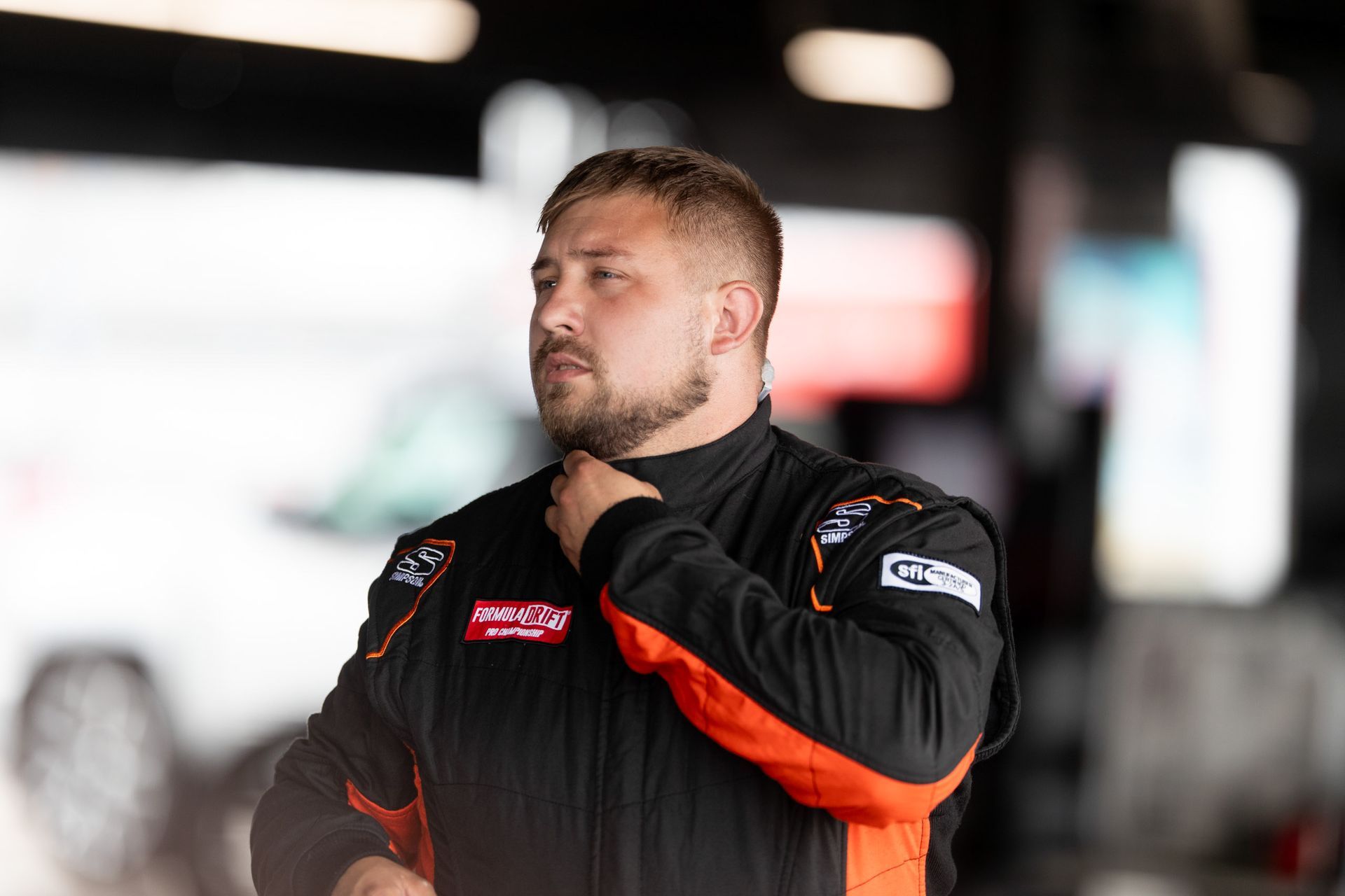 Jonathan Hurst with a beard is wearing a black and orange racing suit.