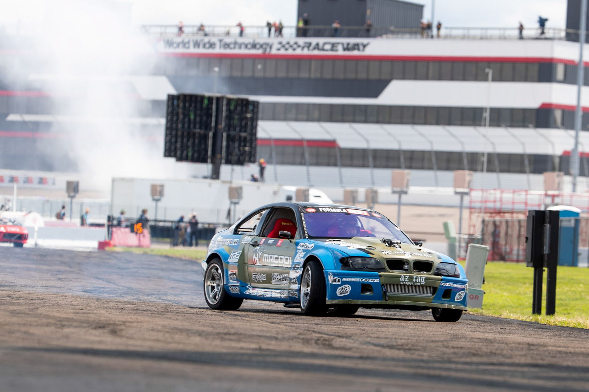 A blue car is driving down a race track in front of a building.